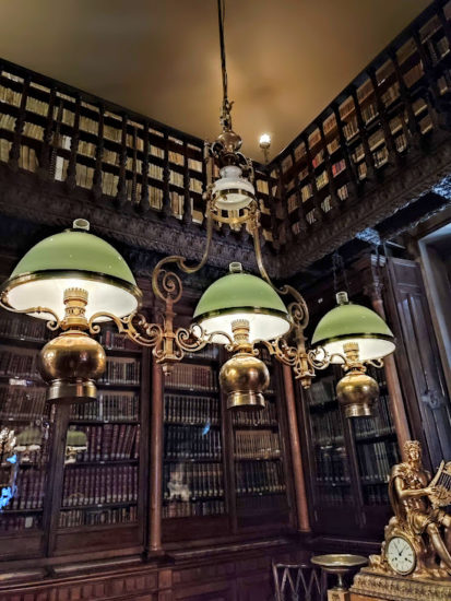 Biblioteca con muchos volúmenes y dos pisos. Decorada con una lámpara de tres brazos.